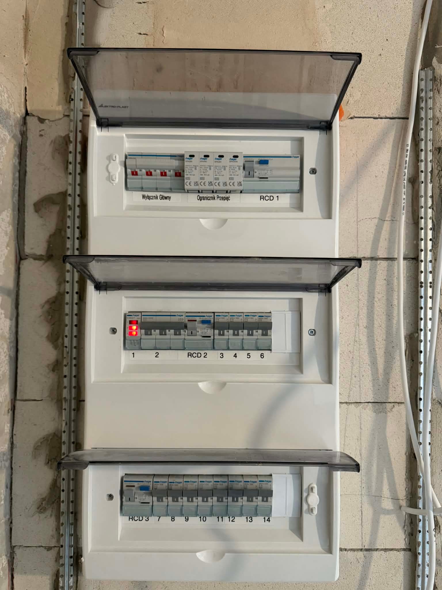 Rozdzielnica elektryczna Hager z otwartymi klapkami, widoczne zabezpieczenia RCD i wyłączniki, zamontowana na ścianie z surowego betonu komórkowego.