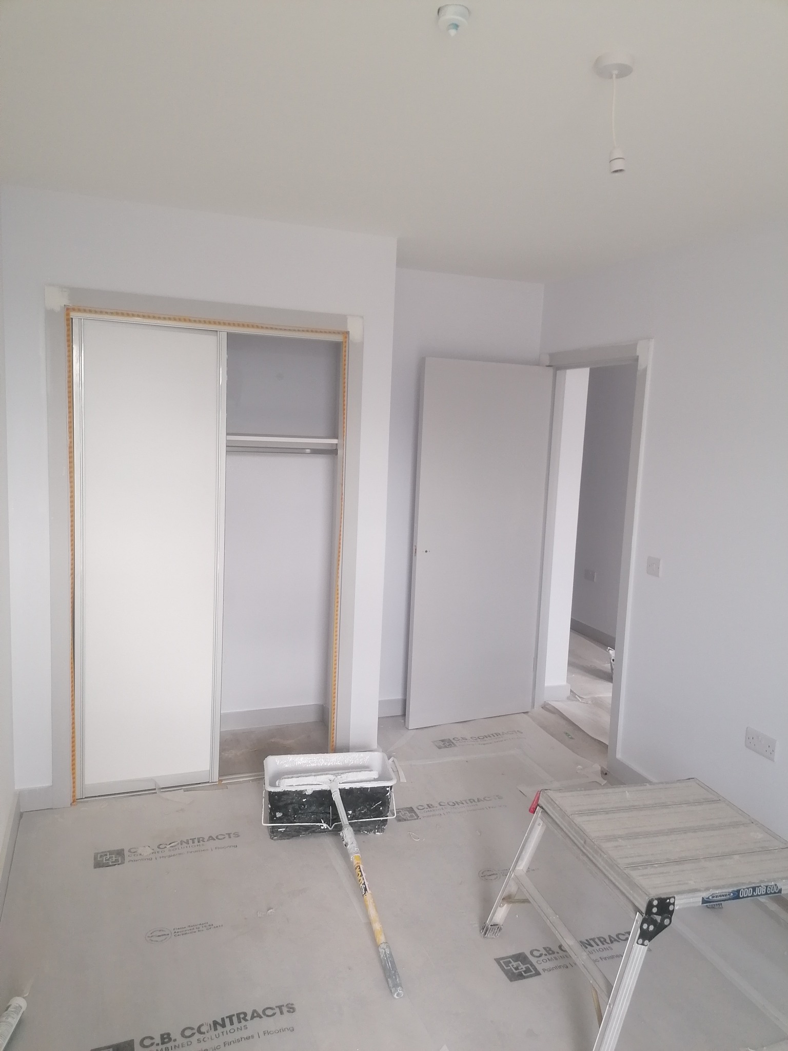 Białe, świeżo pomalowane wnętrze z szafą wnękową i otwartymi drzwiami. Na podłodze folia ochronna, wałek malarski i drabina. Minimalistyczny wystrój.