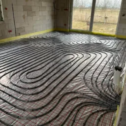 Układanie rur ogrzewania podłogowego na siatce w surowym wnętrzu. Widoczne precyzyjne spirale grzewcze, okna i ściany z białego bloczka. Żółta folia zabezpieczająca.