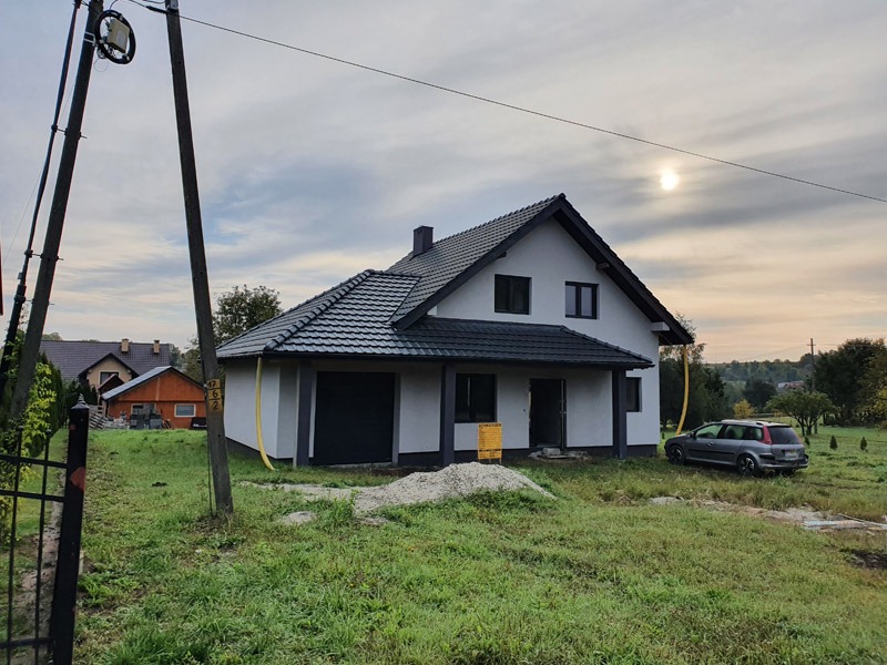 Nowoczesny dom jednorodzinny z garażem dwustanowiskowym w trakcie wykończenia, widok z zewnątrz na tle pochmurnego nieba i zieleni.
