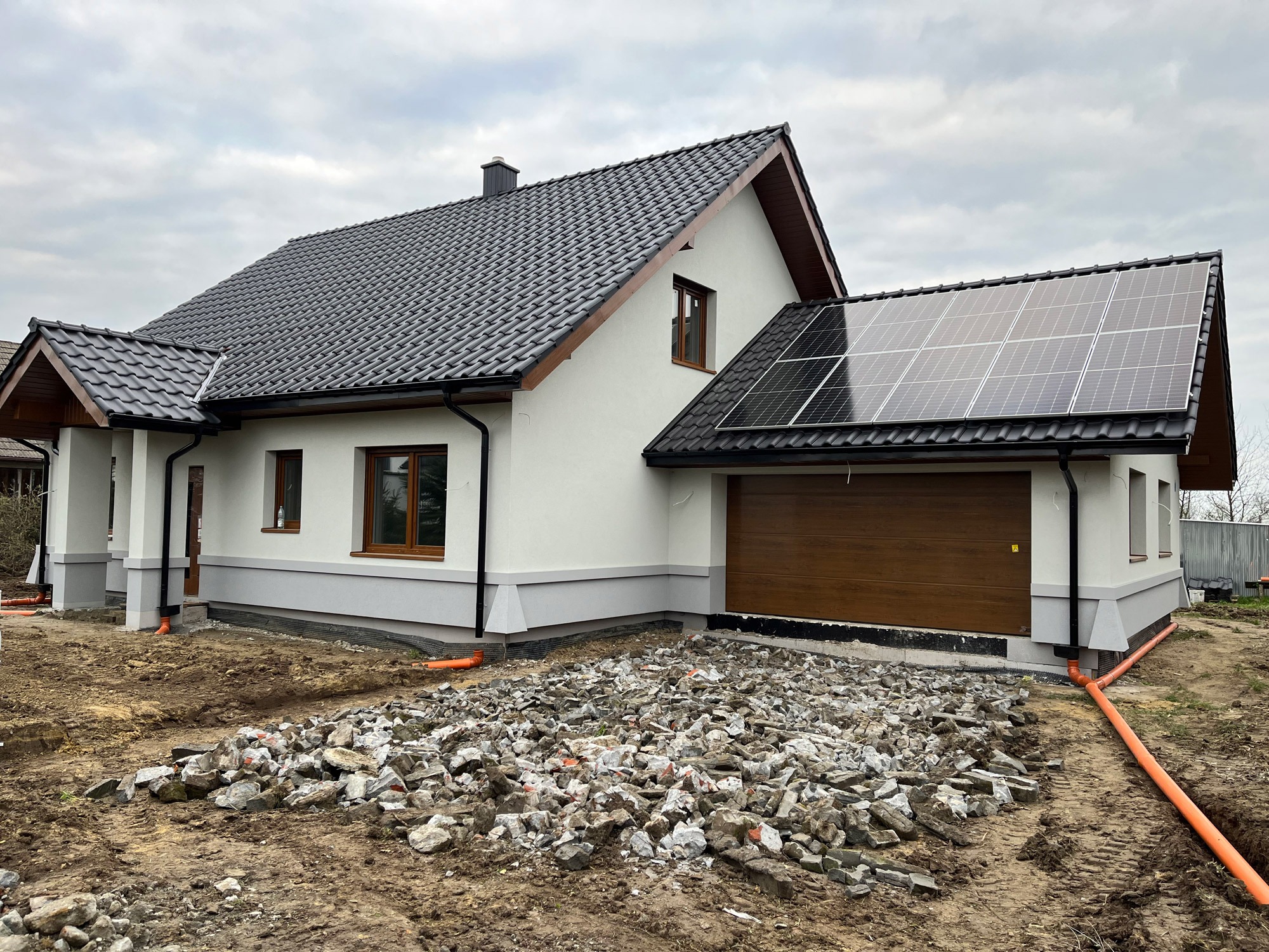 Biały dom z poddaszem i panelami solarnymi na dachu, brązowe okna, gruz przed garażem. Widok ukończonego domu z elementami wykończeniowymi na zewnątrz.