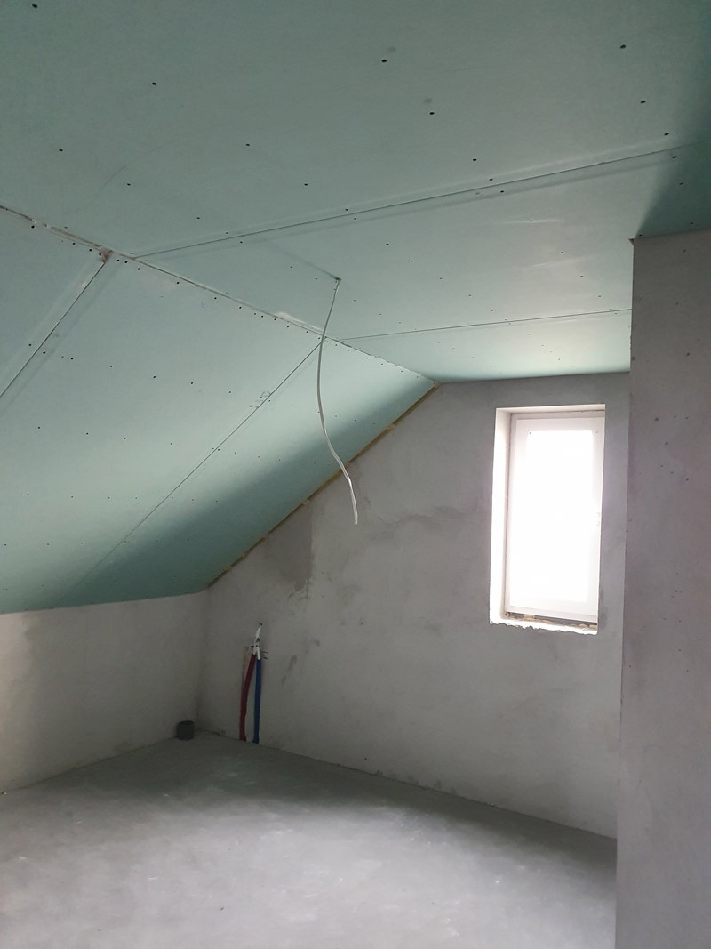 Skośny sufit z płyt gipsowo-kartonowych w pokoju na poddaszu, widok na okno i rury. Ściany surowe, przygotowane do malowania. Etap prac wykończeniowych.