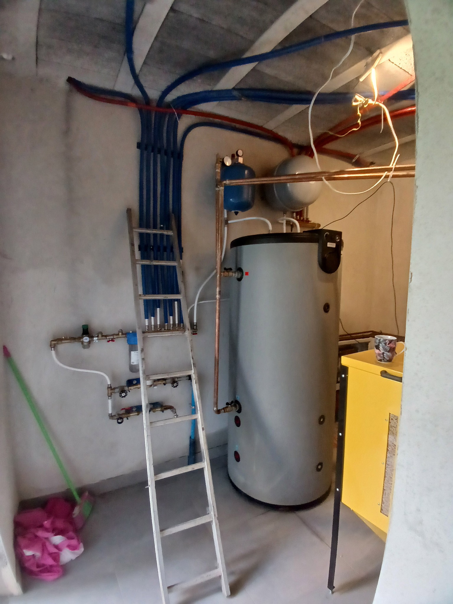 Kotłownia z szarym zbiornikiem, niebieskimi i czerwonymi rurami. Aluminiowa drabina oparta o ścianę. Widoczny żółty piec i instalacja z zaworami.