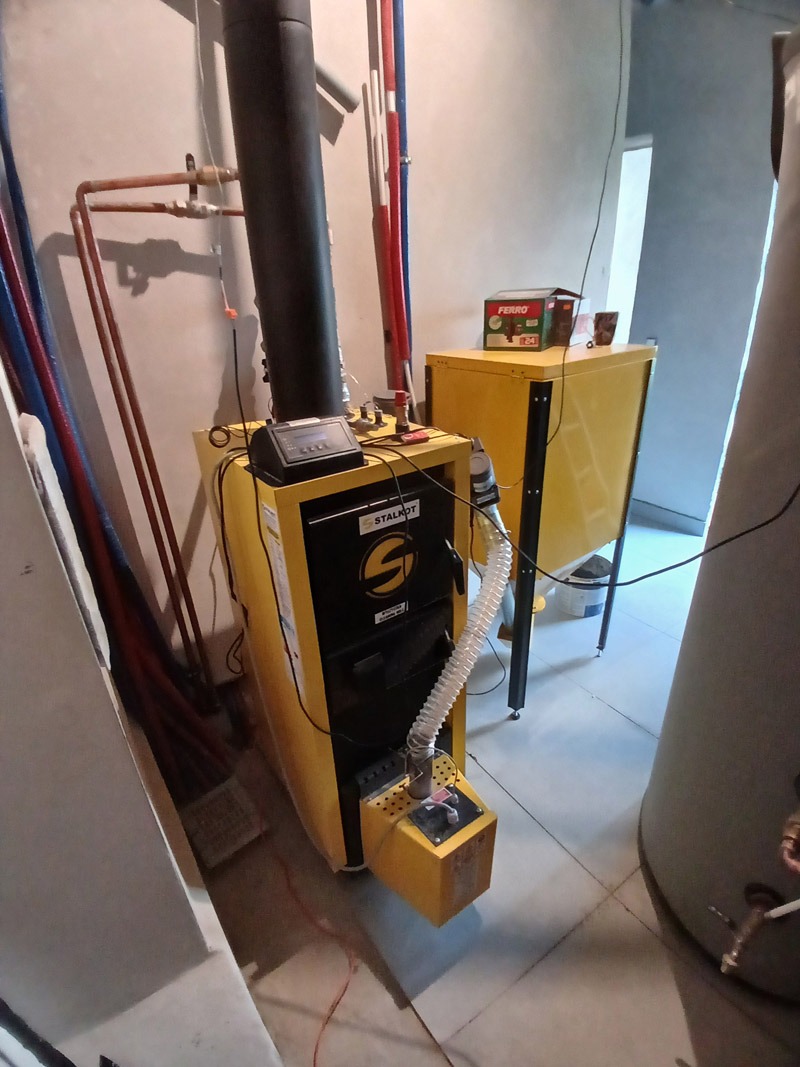 Żółto-czarny kocioł na pellet Stalmark z automatycznym podajnikiem, rury instalacji CO w tle. Ekonomiczne i ekologiczne ogrzewanie.