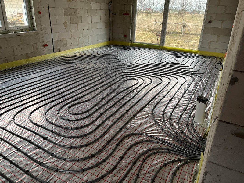 Układanie rur ogrzewania podłogowego na siatce w surowym wnętrzu. Widoczne precyzyjne spirale grzewcze, okna i ściany z białego bloczka. Żółta folia zabezpieczająca.