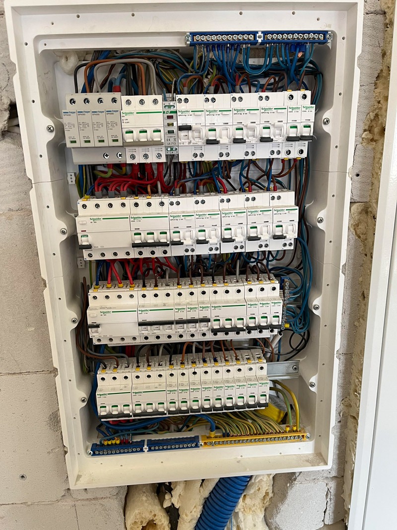 Rozdzielnica elektryczna Schneider w białej obudowie, widoczne zabezpieczenia i kable instalacyjne na tle surowej ściany. Precyzyjny montaż.