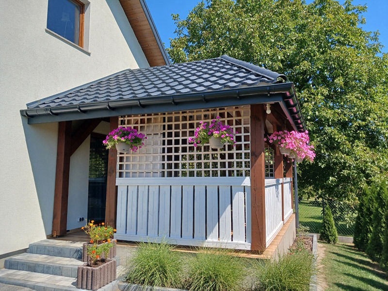 Zadaszone wejście do domu z drewnianą konstrukcją, białymi panelami i kratką. Wiszące donice z różowymi kwiatami dodają uroku. W tle zieleń ogrodu.