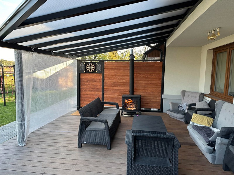 Nowoczesny taras z pergolą: deski Cumaru, aluminiowa konstrukcja z poliwęglanem, strefa relaksu z kominkiem. Na ścianie tarcza do darta.