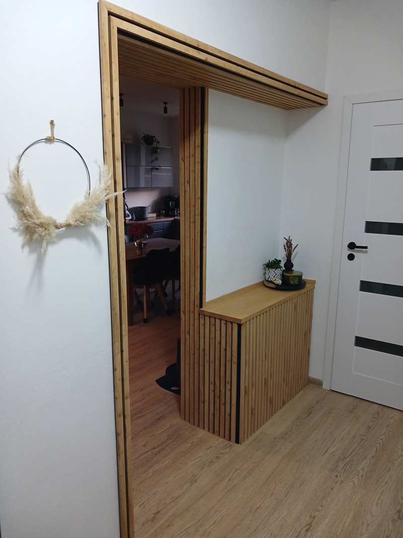 Nowoczesny przedpokój z lamelami: drewniane wykończenie wnęki, minimalistyczny wieniec dekoracyjny, widok na kuchnię. Eleganckie i funkcjonalne rozwiązanie.