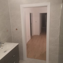 Dariusz Mórawski - Nowoczesna łazienka z szarymi płytkami imitującymi marmur, umywalką z szufladami i białymi drzwiami. Widok na korytarz z drewnianą podłogą.