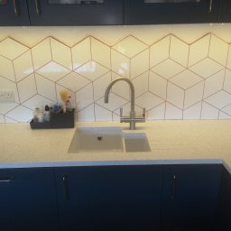 Dariusz Mórawski - Białe, heksagonalne płytki z miedzianymi fugami nad zlewem w kuchni. Blat z jasnego kamienia, ciemne szafki. Nowoczesne wykończenie, oświetlenie LED.