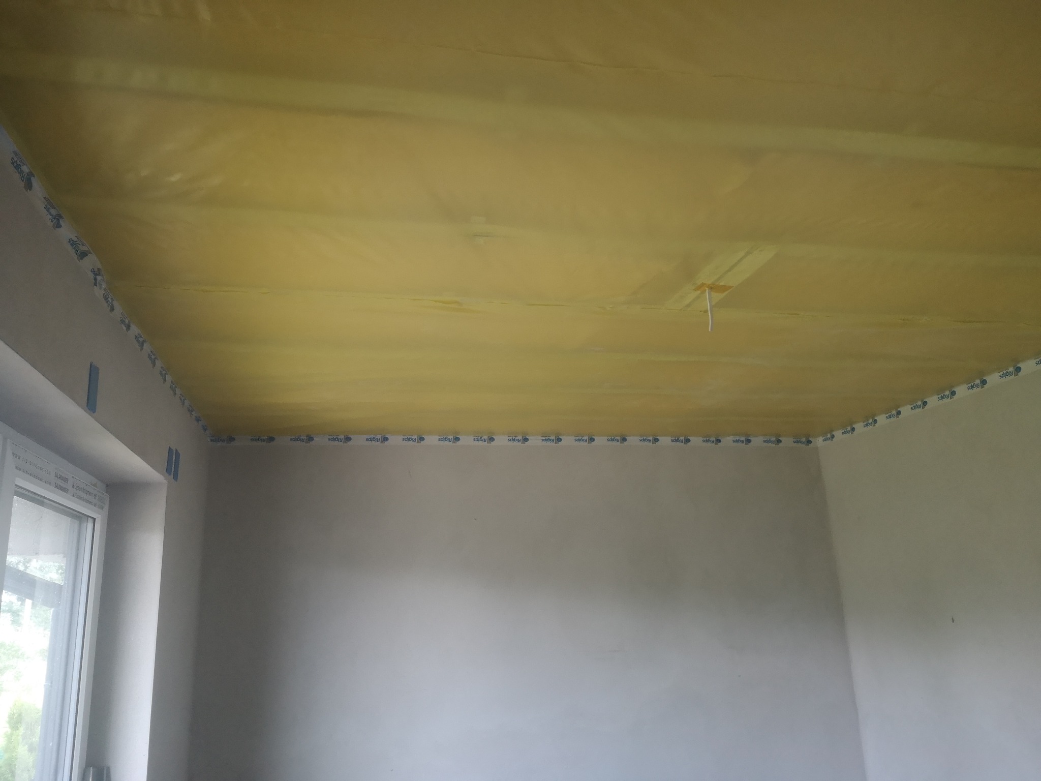 Przygotowanie sufitu do prac wykończeniowych: żółta folia paroizolacyjna zamocowana taśmą w pomieszczeniu z oknem i surowymi ścianami.