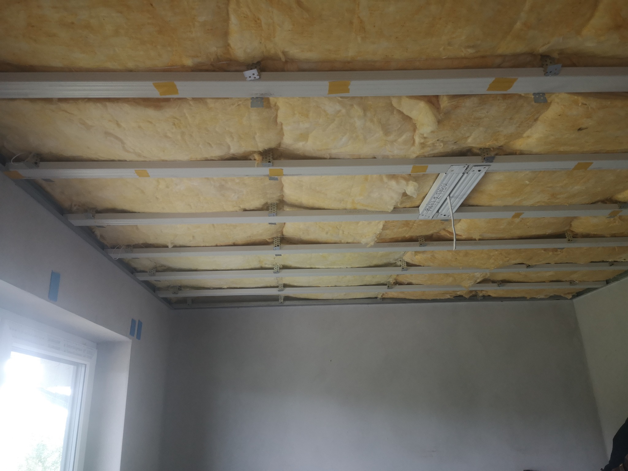 Ocieplenie poddasza wełną mineralną, widoczne metalowe profile mocujące. Fragment okna i surowa ściana w tle. Prace wykończeniowe w trakcie realizacji.