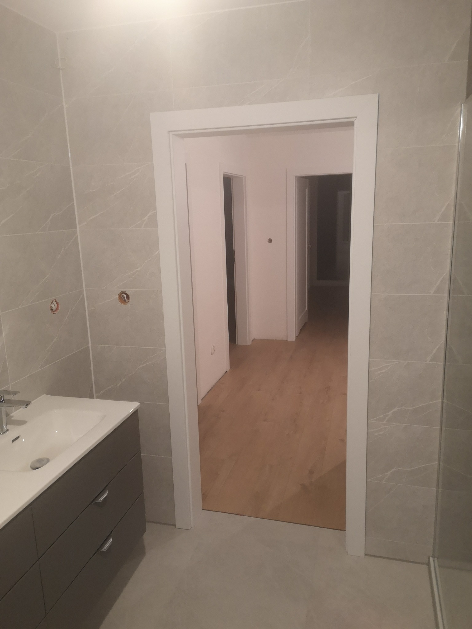 Nowoczesna łazienka z szarymi płytkami imitującymi marmur, umywalką z szufladami i białymi drzwiami. Widok na korytarz z drewnianą podłogą.