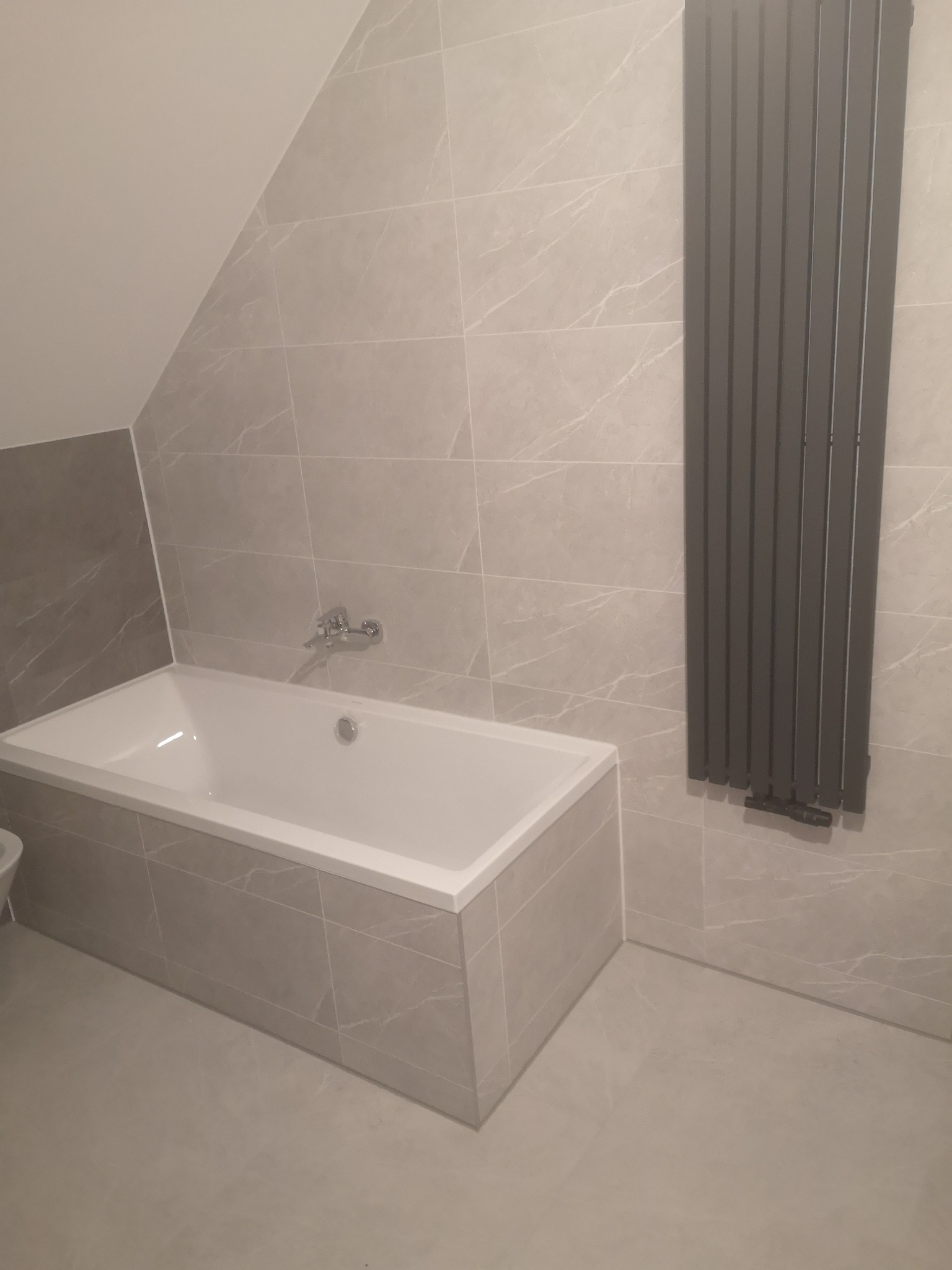 Wykończona łazienka na poddaszu z wanną obudowaną płytkami imitującymi marmur i szarym grzejnikiem drabinkowym na ścianie z jasnych płytek.