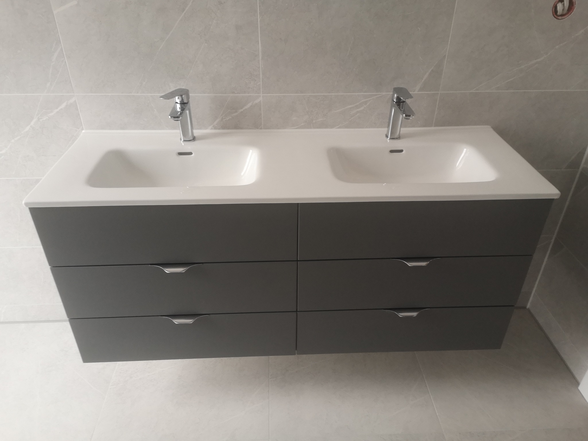 Nowoczesna, szara szafka pod umywalkę z dwiema białymi umywalkami i chromowanymi bateriami na tle płytek imitujących marmur. Minimalistyczny design.