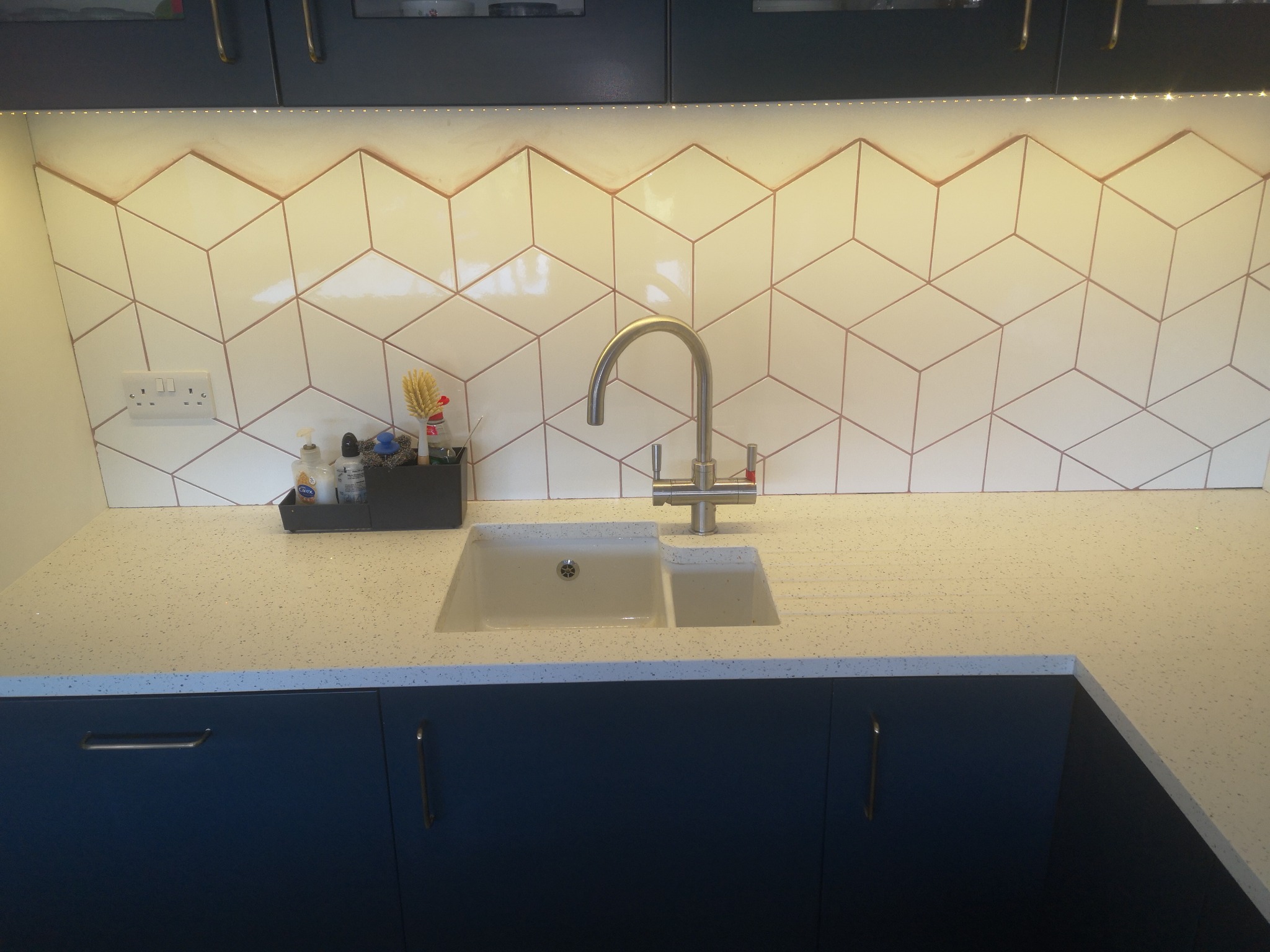 Białe, heksagonalne płytki z miedzianymi fugami nad zlewem w kuchni. Blat z jasnego kamienia, ciemne szafki. Nowoczesne wykończenie, oświetlenie LED.
