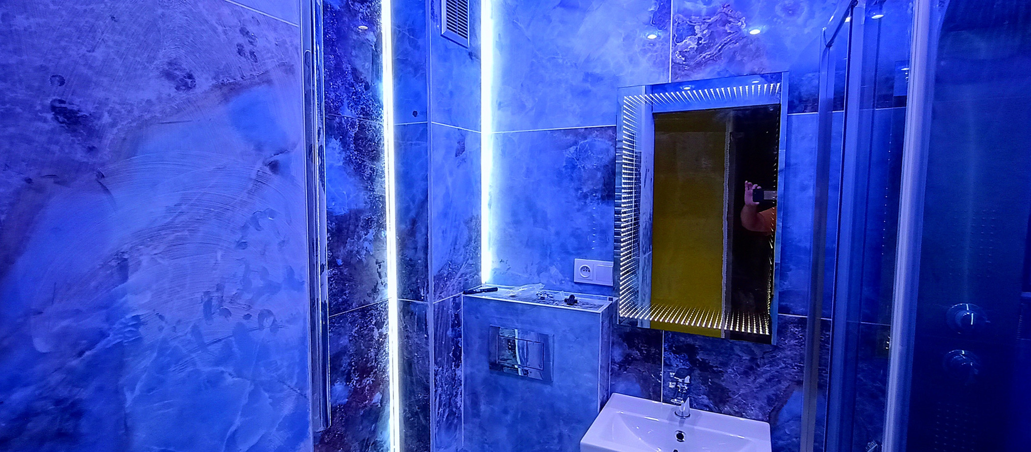 Niebieska łazienka z marmurowymi ścianami, podświetlanymi listwami LED, lustrem z efektem nieskończoności i kabiną prysznicową. Nowoczesny design wnętrza.