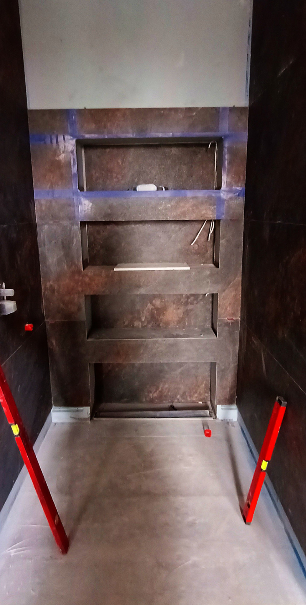 Nisze w ścianie z ciemnej płytki, zabezpieczone taśmą malarską, poziomice na podłodze. Etap prac wykończeniowych w łazience.