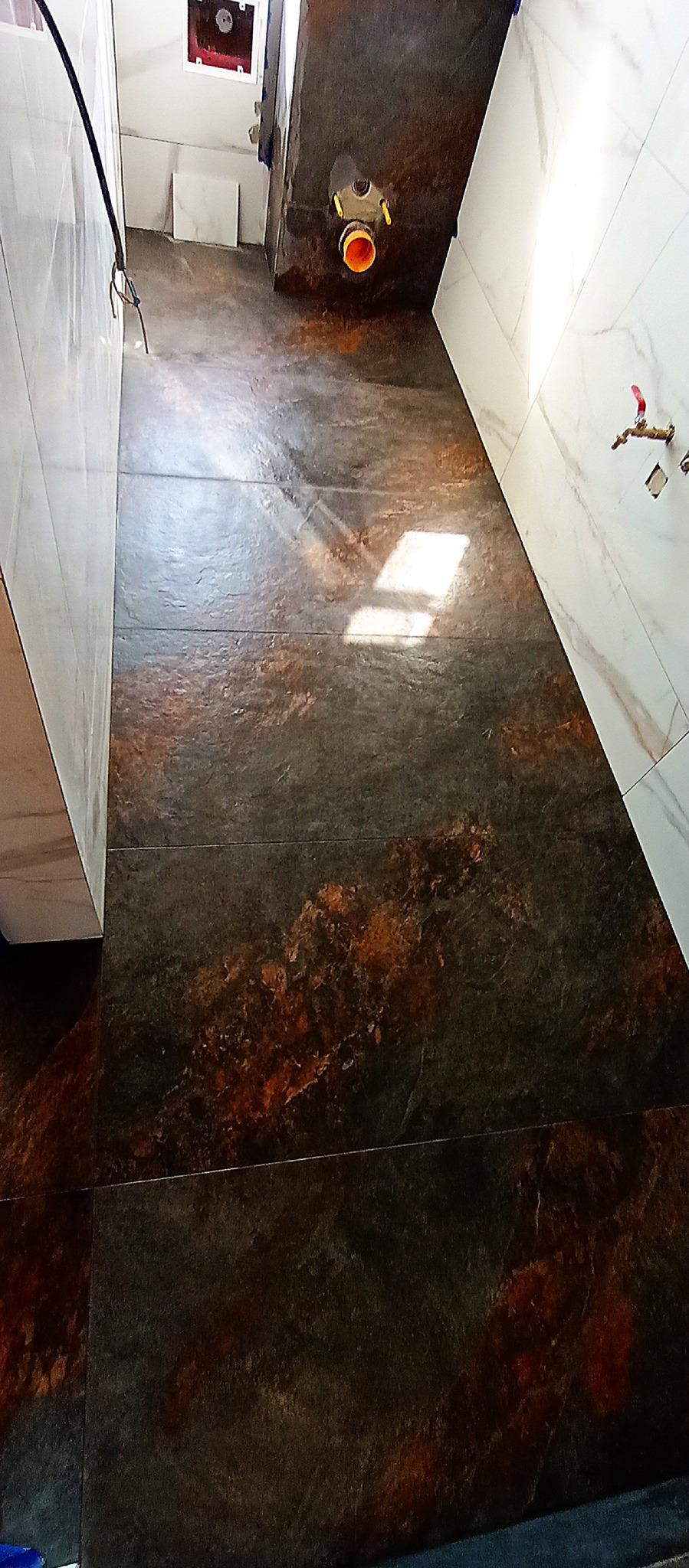 Wykończona łazienka z ciemnymi, wzorzystymi płytkami podłogowymi i jasnymi płytkami ściennymi. Widoczny odpływ liniowy i fragment instalacji.