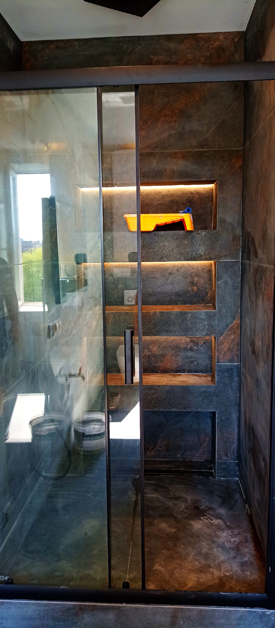 Nowoczesna kabina prysznicowa z ciemnymi, kamiennymi płytkami i wnękami z podświetleniem. Widoczne akcesoria malarskie i detale wykończeniowe.