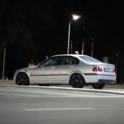Srebrne BMW E46 na tle nocnego krajobrazu miejskiego, oświetlone uliczną lampą. Widoczny bok i tył auta, czarne felgi. Ujęcie z poziomu jezdni.