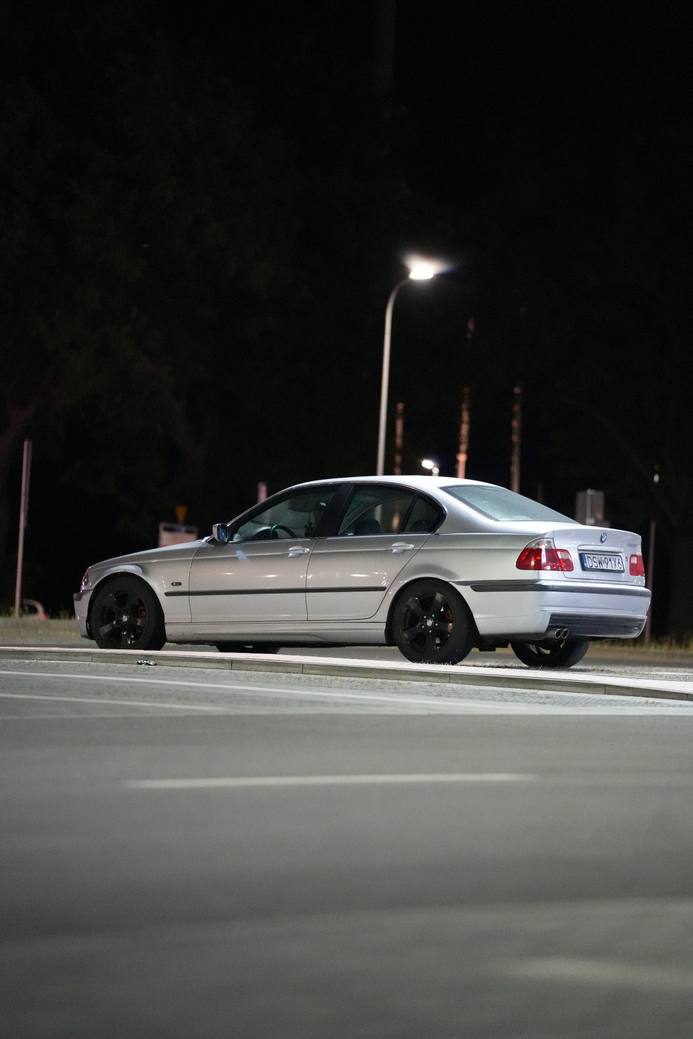 Srebrne BMW E46 na tle nocnego krajobrazu miejskiego, oświetlone uliczną lampą. Widoczny bok i tył auta, czarne felgi. Ujęcie z poziomu jezdni.