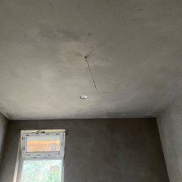Slawbud - Pokój po nałożeniu tynku maszynowego, widoczne ściany i sufit w szarym kolorze. W tle okno z widokiem na zieleń. W suficie widoczne miejsce na lampę.