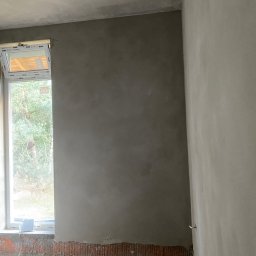 Slawbud - Ściana po nałożeniu tynku w szarym odcieniu, widoczna przy oknie. Częściowo otynkowana ściana z cegieł. Widok na zewnątrz przez okno.