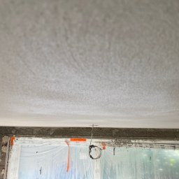 Slawbud - Świeżo położona, surowa gładź na suficie w trakcie wysychania, widok z dołu. Okno zabezpieczone folią malarską. W tle zarys zieleni za oknem.