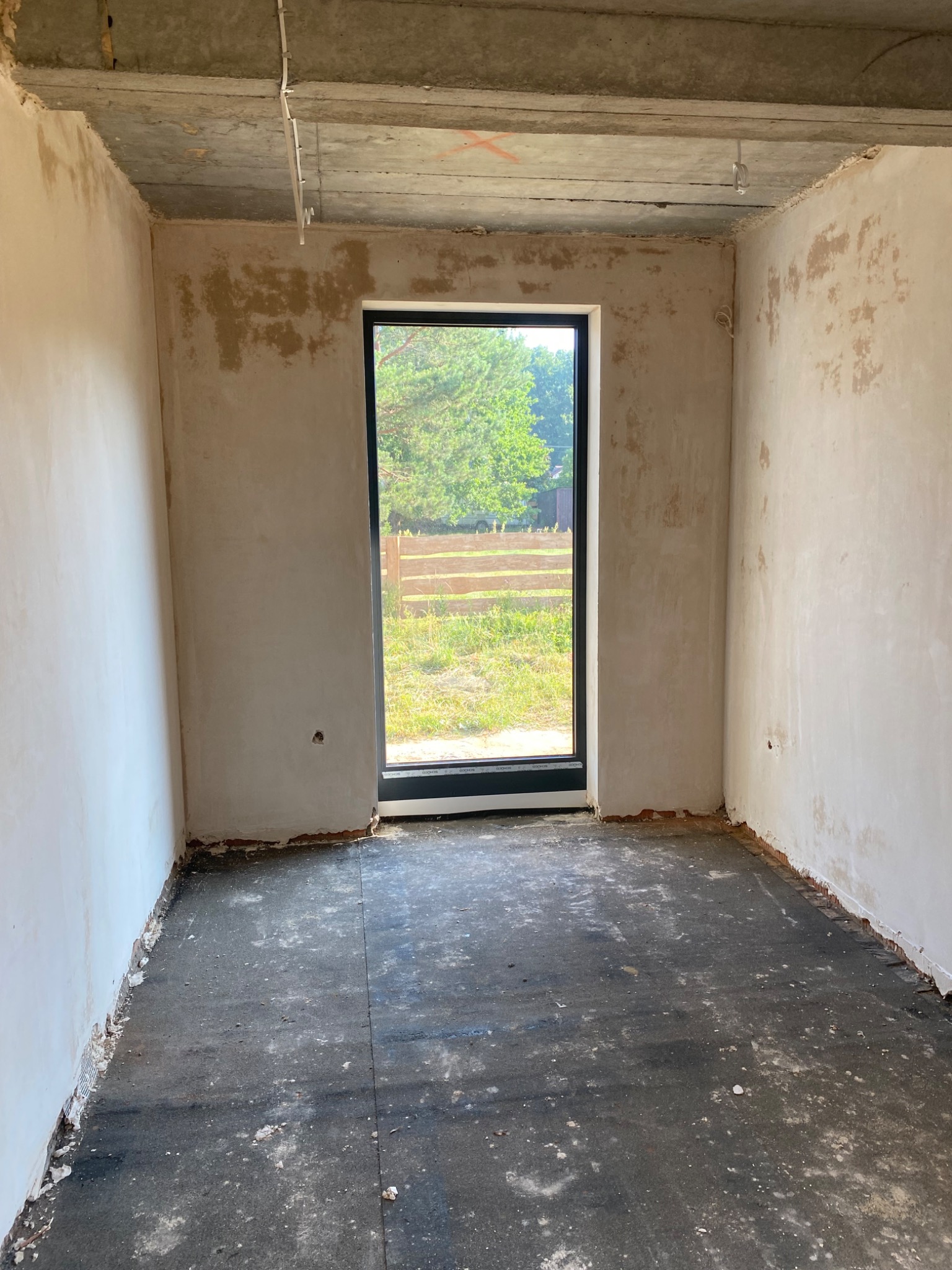 Surowe wnętrze pokoju z oknem, ściany w trakcie tynkowania/szpachlowania, widoczne niedoskonałości i surowy beton na suficie. W tle zieleń za oknem.