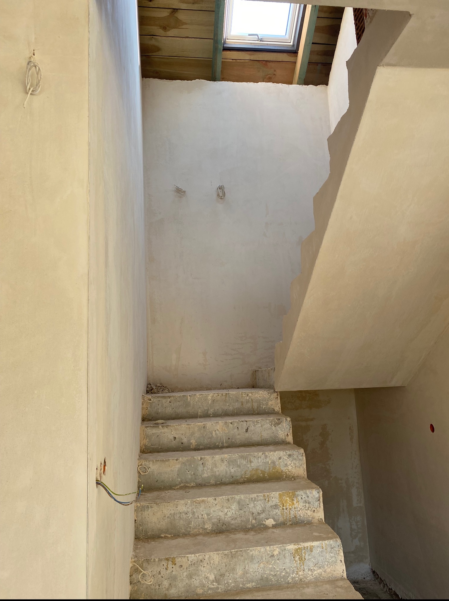 Wnętrze w trakcie prac wykończeniowych: ściany i schody pokryte świeżym tynkiem, widoczne przewody elektryczne i okno dachowe. Surowy stan, gotowy do dalszych prac.