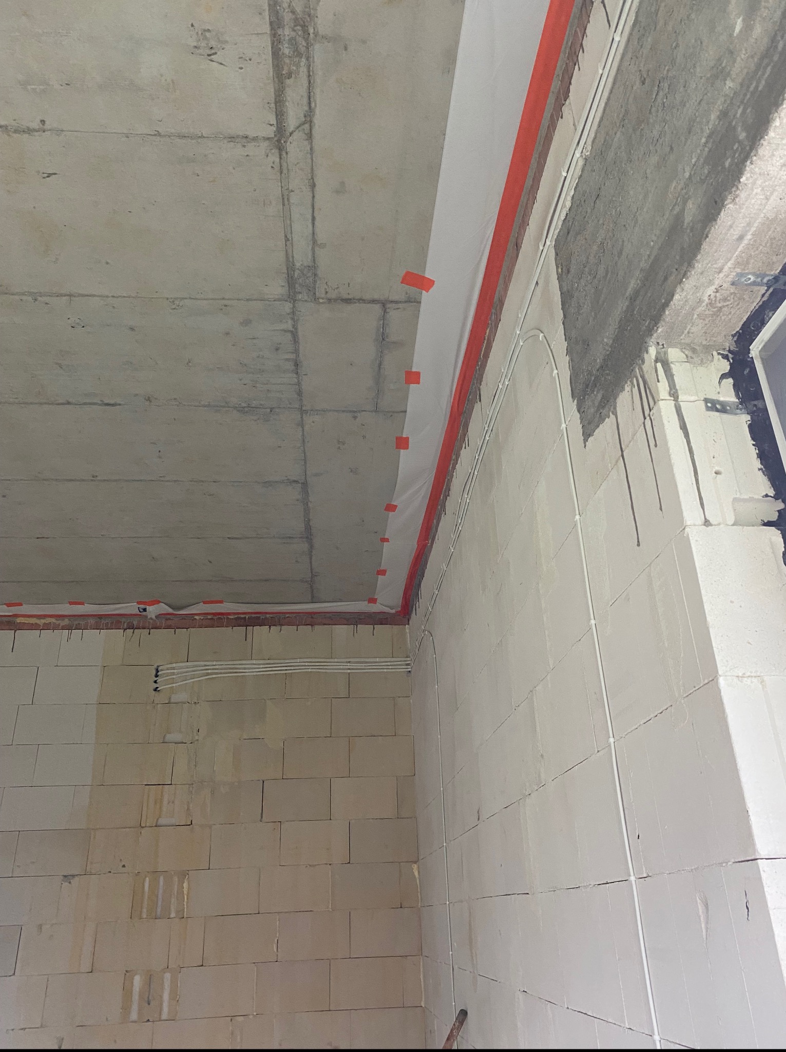 Surowy stan: ściana z pustaków, betonowy sufit, instalacja elektryczna w białych rurkach, zabezpieczona folią malarską z pomarańczową taśmą.  Wnętrze w trakcie prac.