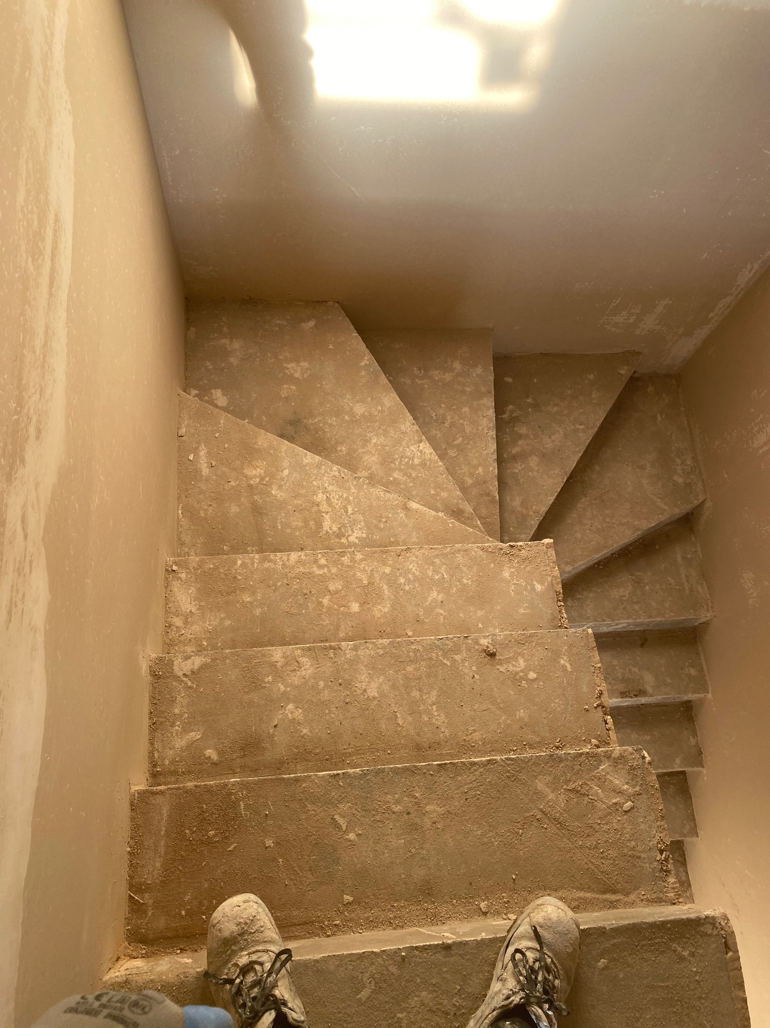 Wnętrze w trakcie prac wykończeniowych: schody i ściany pokryte świeżym tynkiem. Widoczne buty pracownika, co sugeruje trwający remont.