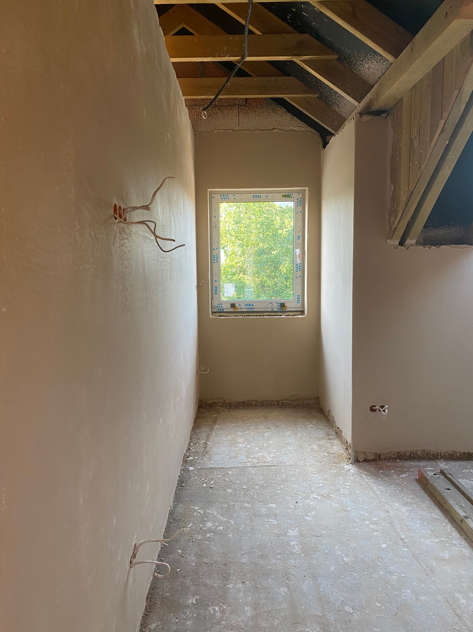 Wnętrze pokoju po nałożeniu tynku, widoczne wystające kable elektryczne, okno z widokiem na zieleń oraz drewniana konstrukcja dachu. Ściany w jasnym odcieniu, surowy stan podłogi.