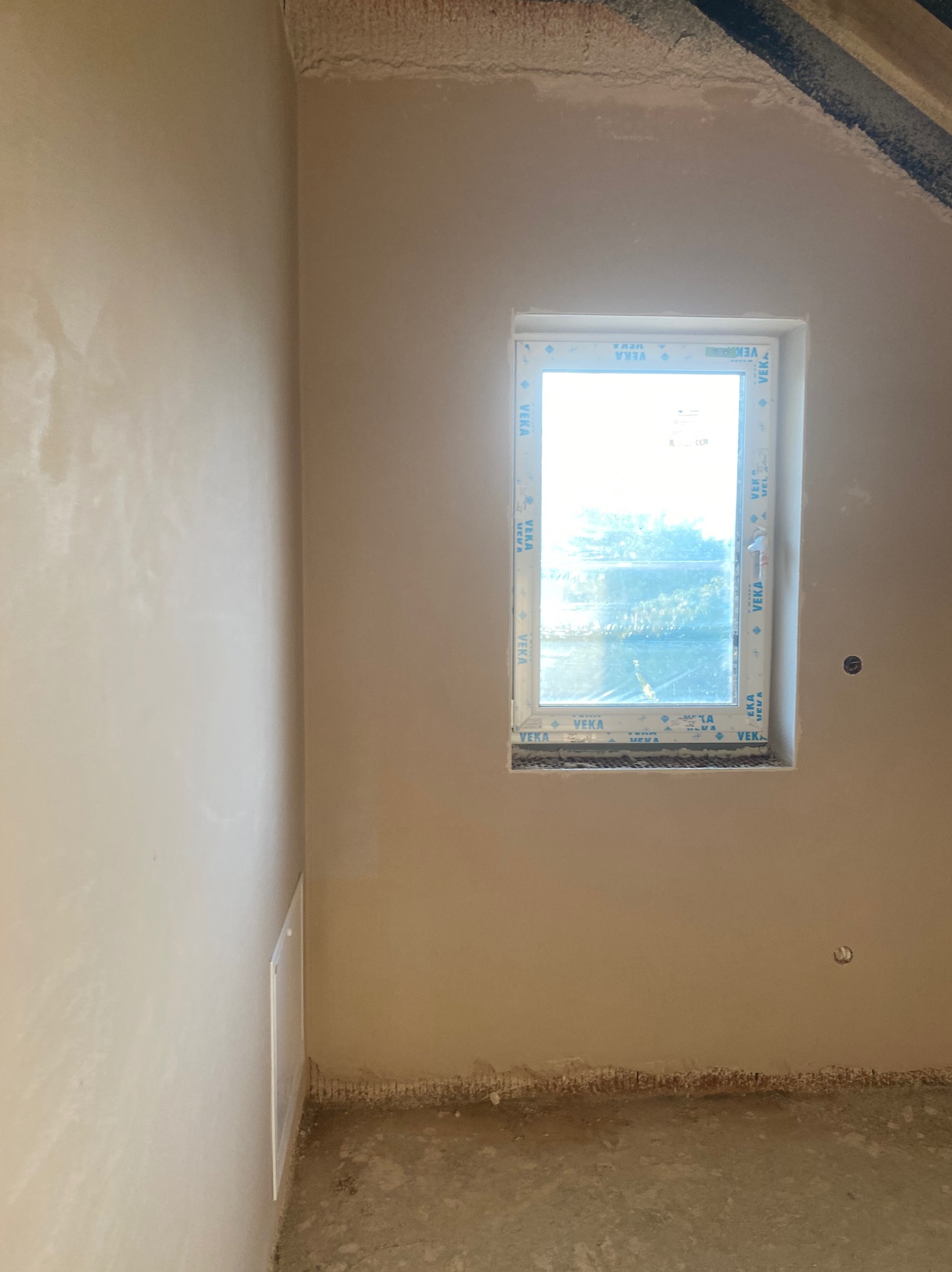 Ściana po nałożeniu tynku w pomieszczeniu z oknem Veka. Widoczne nierówności i przygotowanie do dalszych prac wykończeniowych. Na dole ściany widoczne pozostałości po pracach.