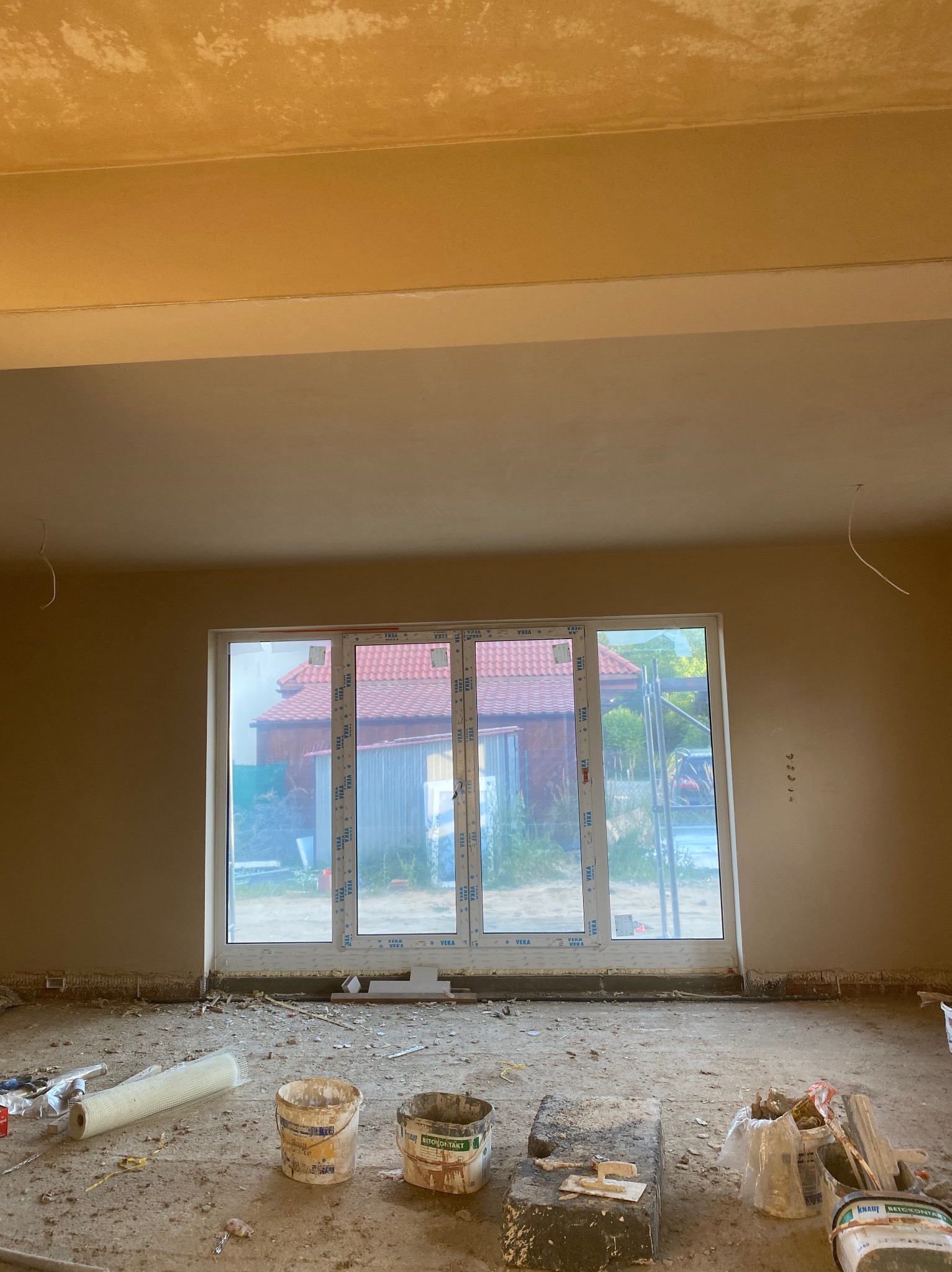 Pokój w trakcie prac wykończeniowych: ściany przygotowane do malowania, widoczne narzędzia i materiały budowlane. Okno z widokiem na zewnątrz.