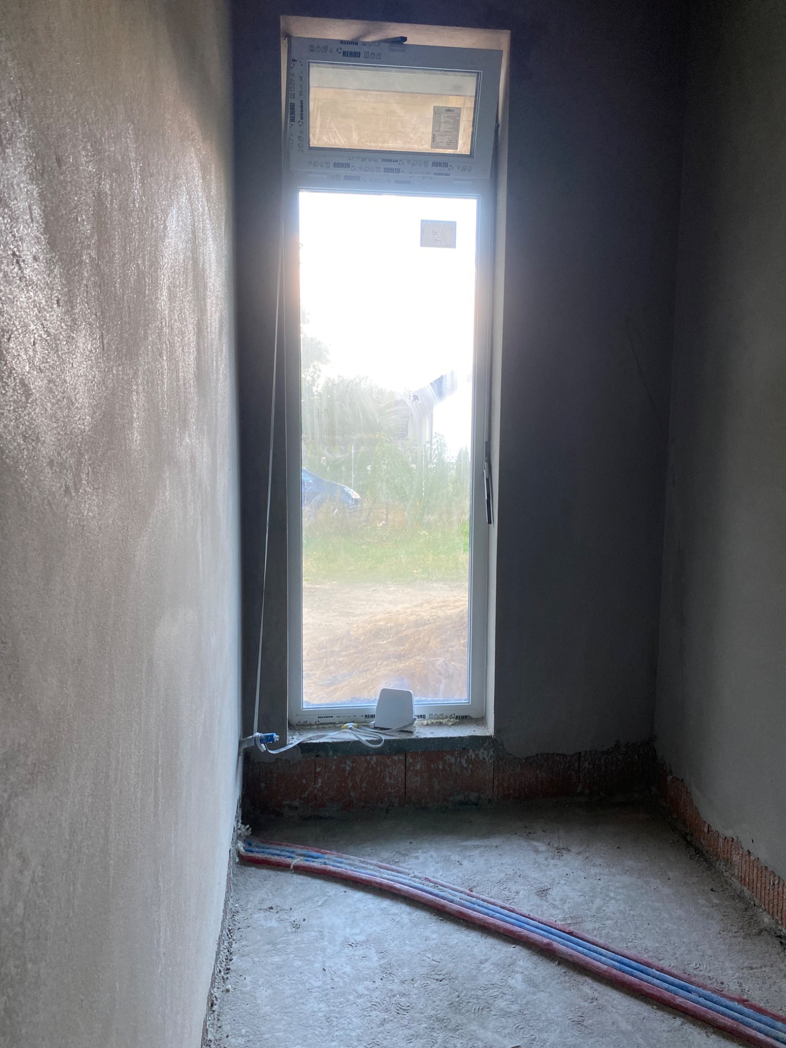 Surowe wnętrze z oknem i instalacją. Ściany w trakcie prac wykończeniowych, widoczne ślady po tynkowaniu i instalacje podłogowe. Oświetlenie dzienne przez okno.