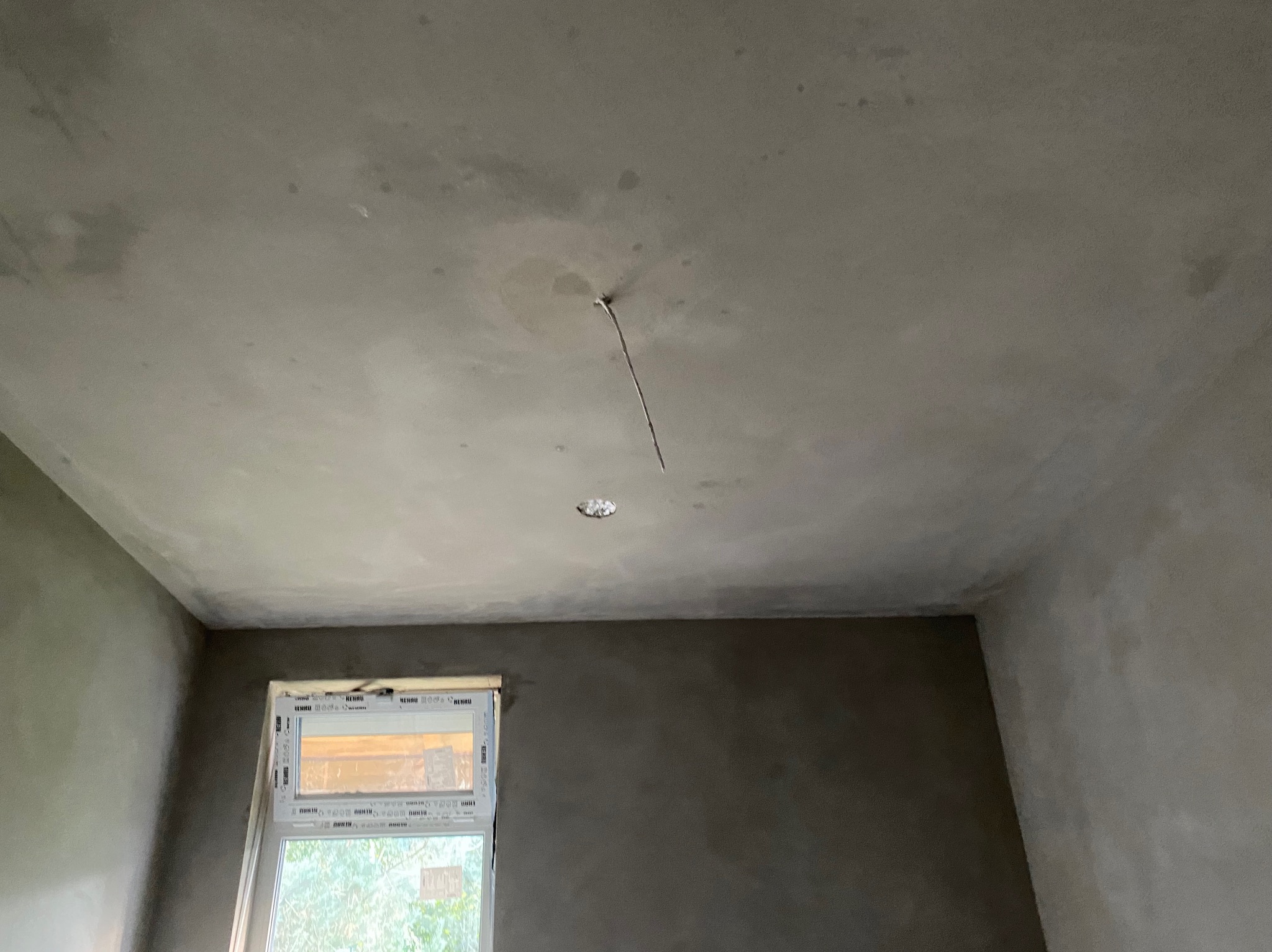 Pokój po nałożeniu tynku maszynowego, widoczne ściany i sufit w szarym kolorze. W tle okno z widokiem na zieleń. W suficie widoczne miejsce na lampę.