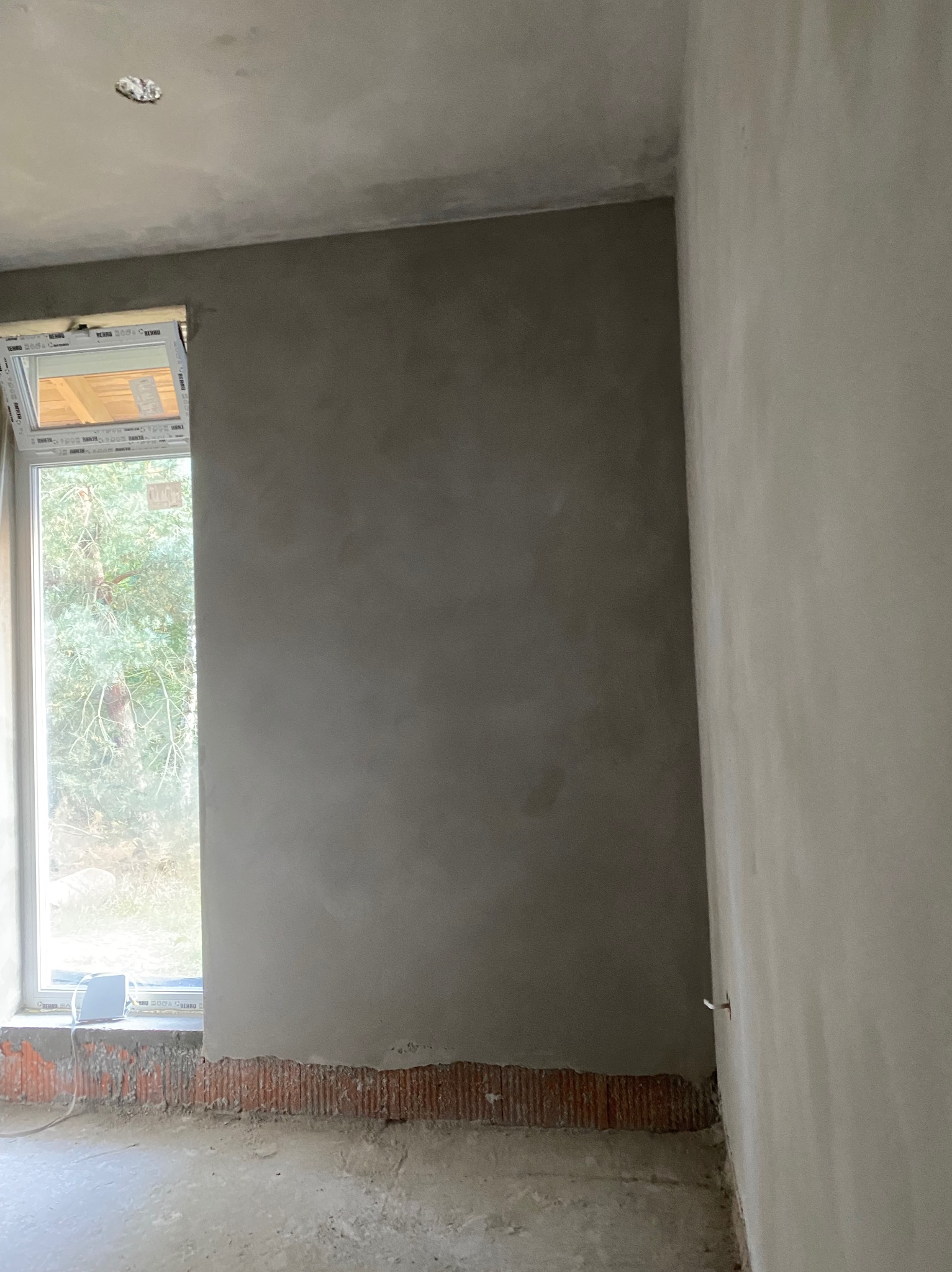 Ściana po nałożeniu tynku w szarym odcieniu, widoczna przy oknie. Częściowo otynkowana ściana z cegieł. Widok na zewnątrz przez okno.