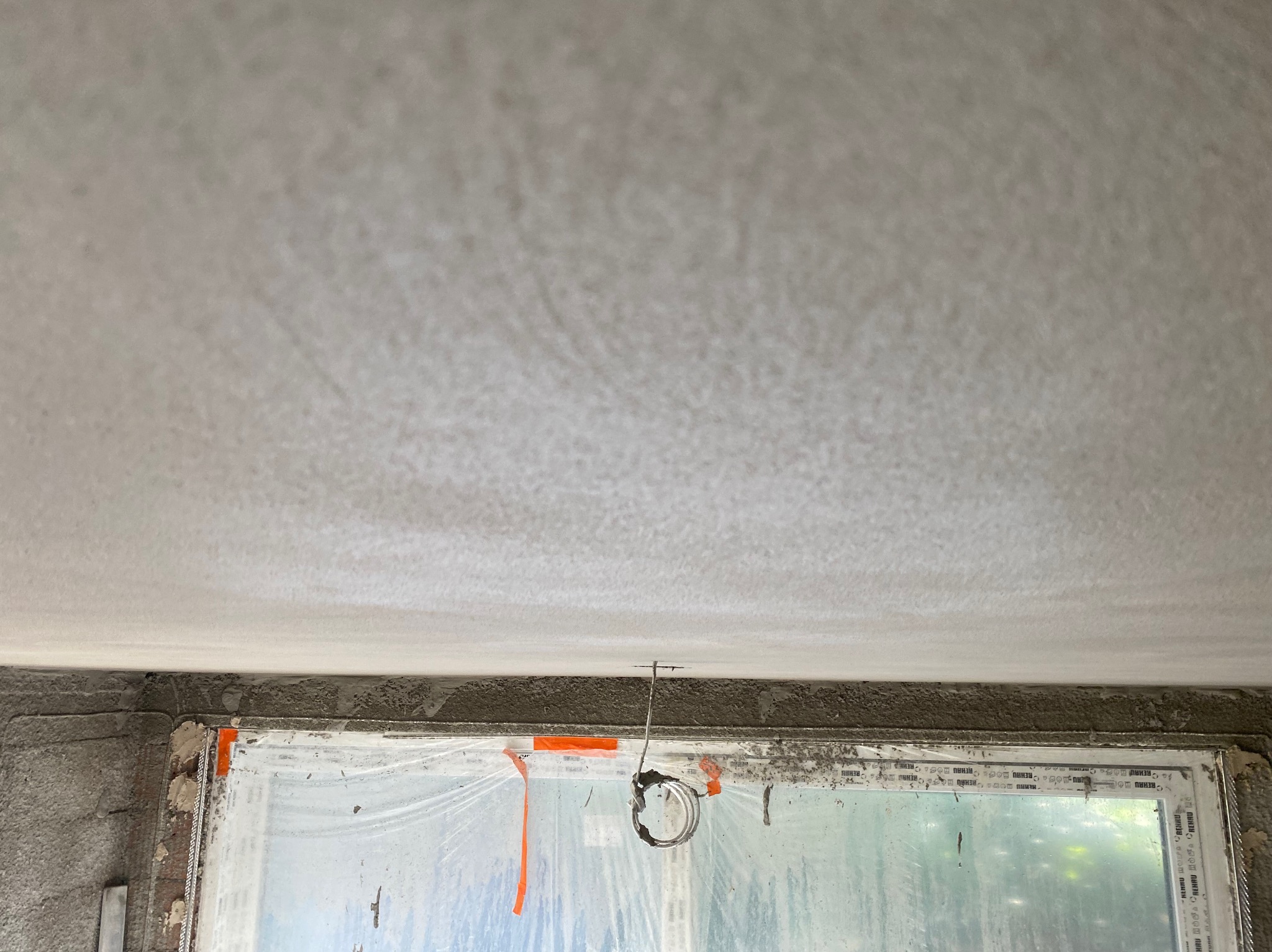 Świeżo położona, surowa gładź na suficie w trakcie wysychania, widok z dołu. Okno zabezpieczone folią malarską. W tle zarys zieleni za oknem.