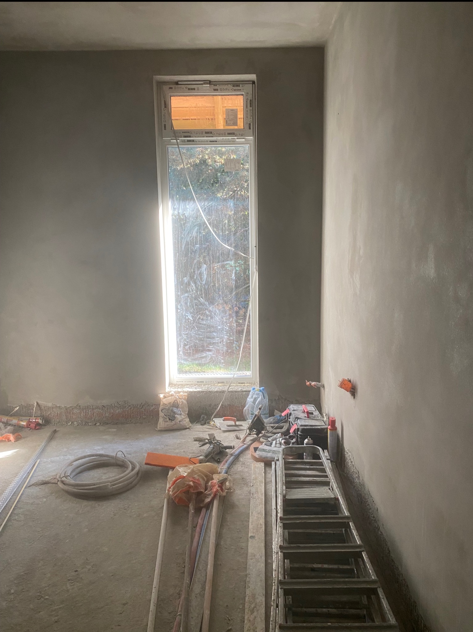 Wnętrze w trakcie prac wykończeniowych: ściany pokryte świeżym tynkiem, okno pionowe, narzędzia i drabina na podłodze. Widoczne ślady prac murarskich.