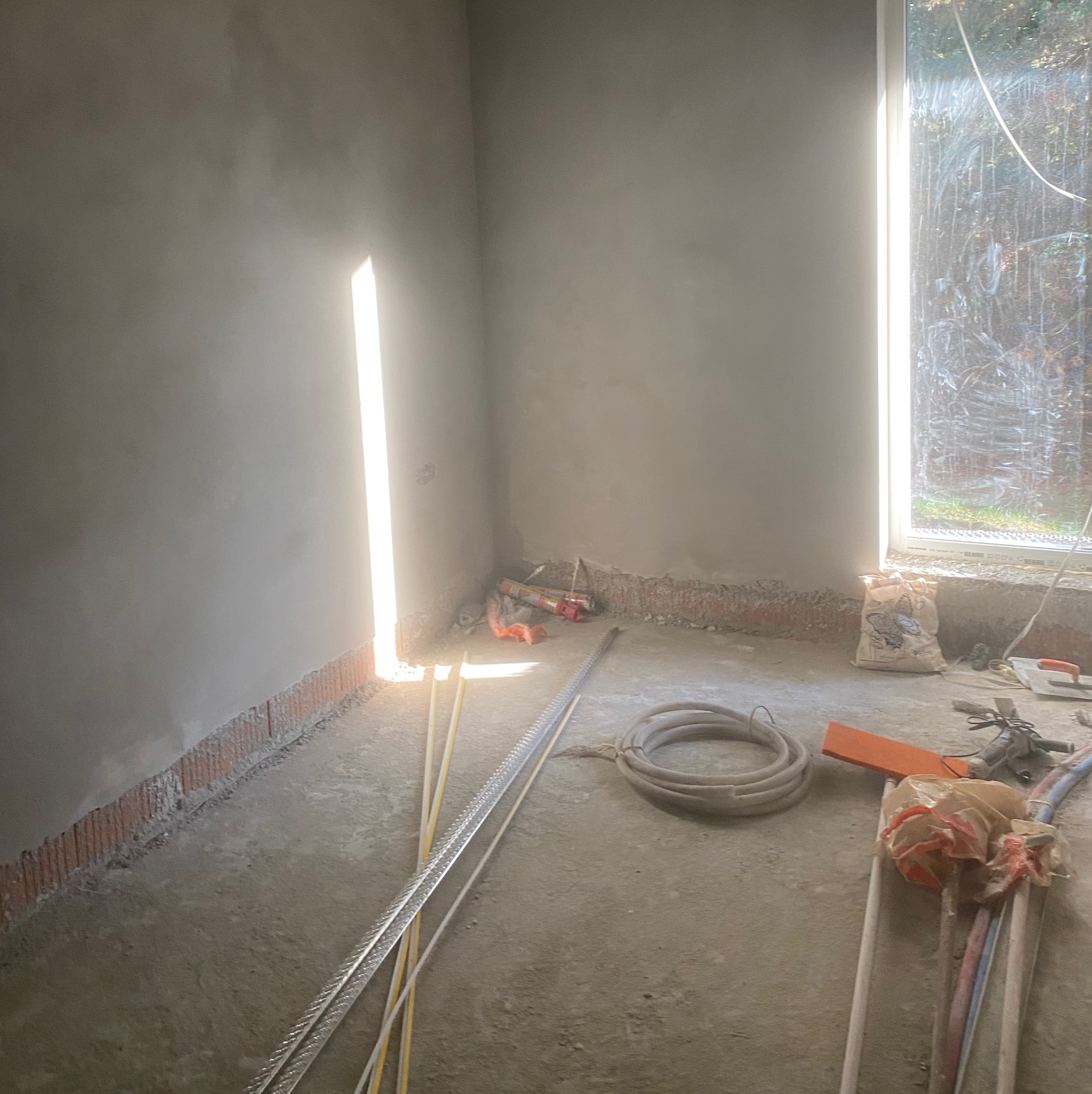 Surowe wnętrze po tynkowaniu ścian, widoczne narzędzia i materiały budowlane, wpadające światło przez okno i szczelinę w ścianie.