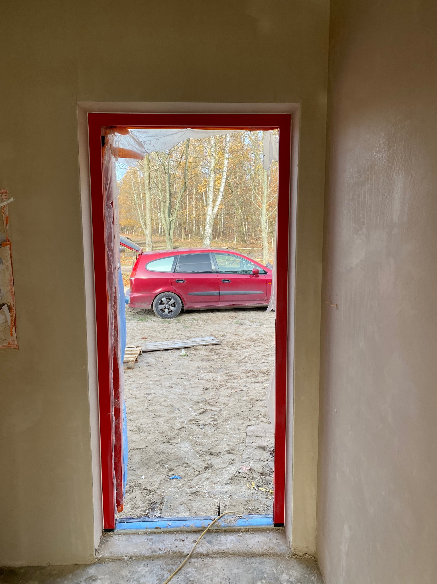 Ściana po tynkowaniu, widok przez czerwone futryny na podwórko z czerwonym autem z otwartym bagażnikiem. Świeże tynki w jasnym odcieniu beżu.