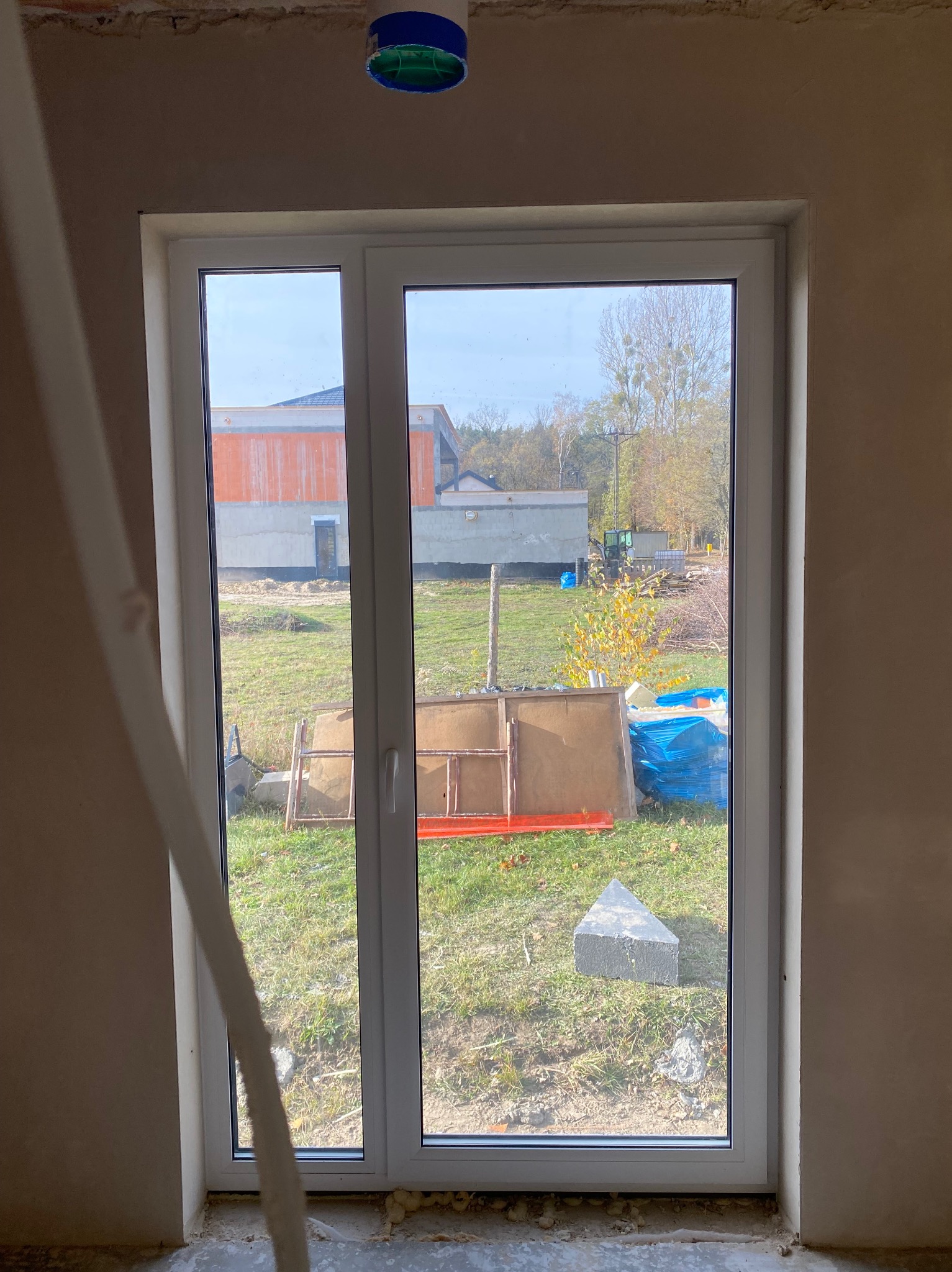 Nowe okno PCV w surowym wnętrzu, ściany przygotowane do wykończenia. Widok na zewnątrz z elementami budowlanymi na trawie. Niebieska puszka elektryczna na suficie.