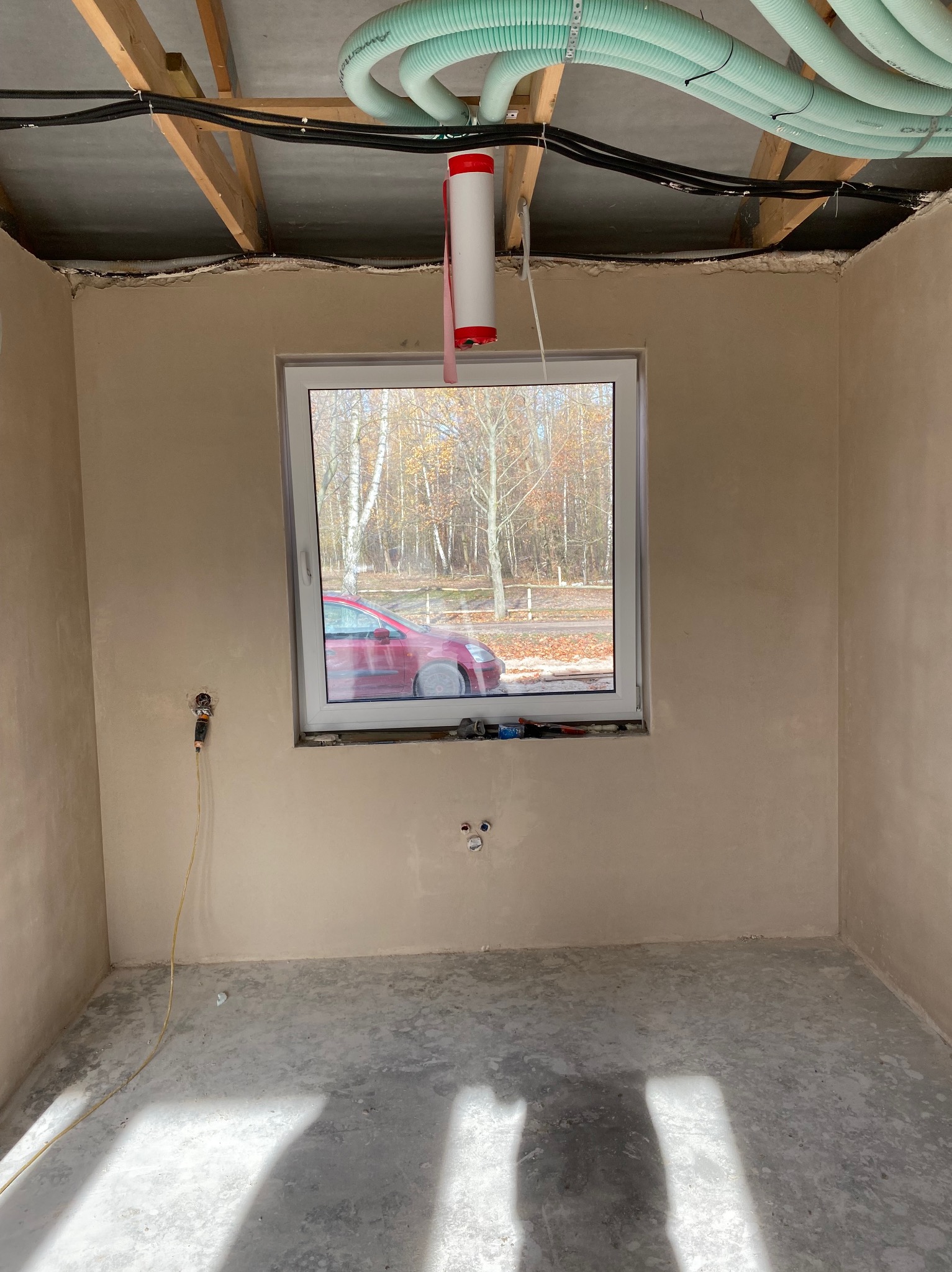 Wnętrze w stanie deweloperskim po tynkowaniu ścian. Widoczne instalacje pod sufitem i okno z widokiem na drzewa. Na parapecie leżą narzędzia.