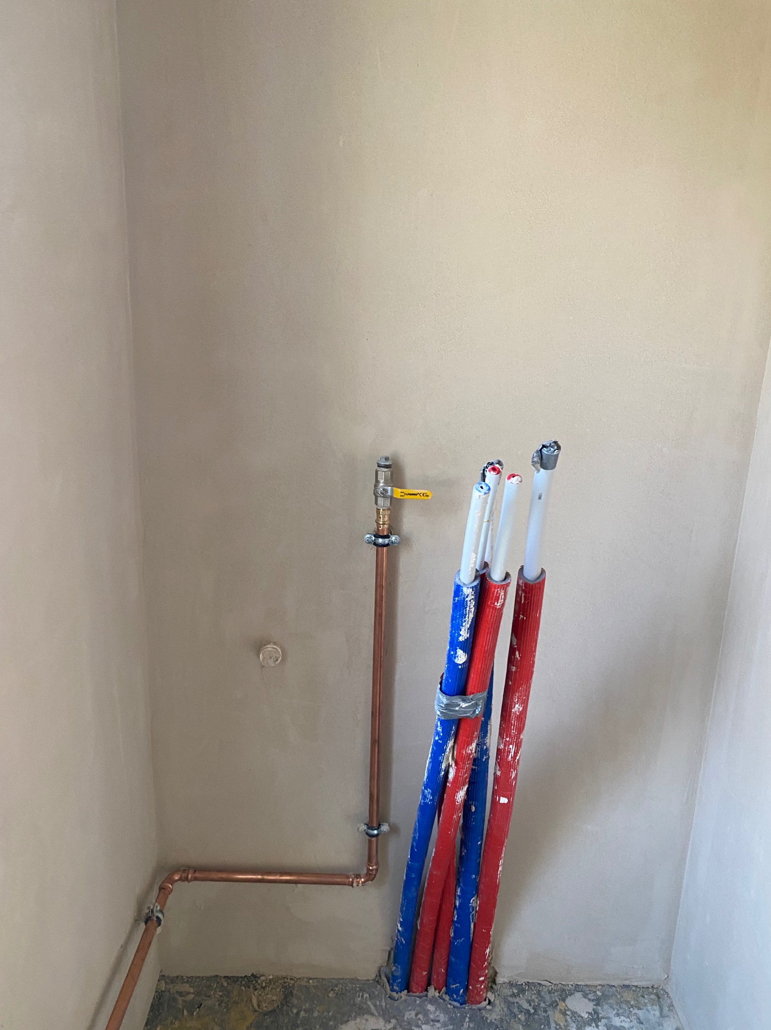 Ściana po szpachlowaniu z instalacją wodną z miedzianych rur i rurami grzewczymi w izolacji czerwono-niebieskiej, przygotowana do dalszych prac wykończeniowych.