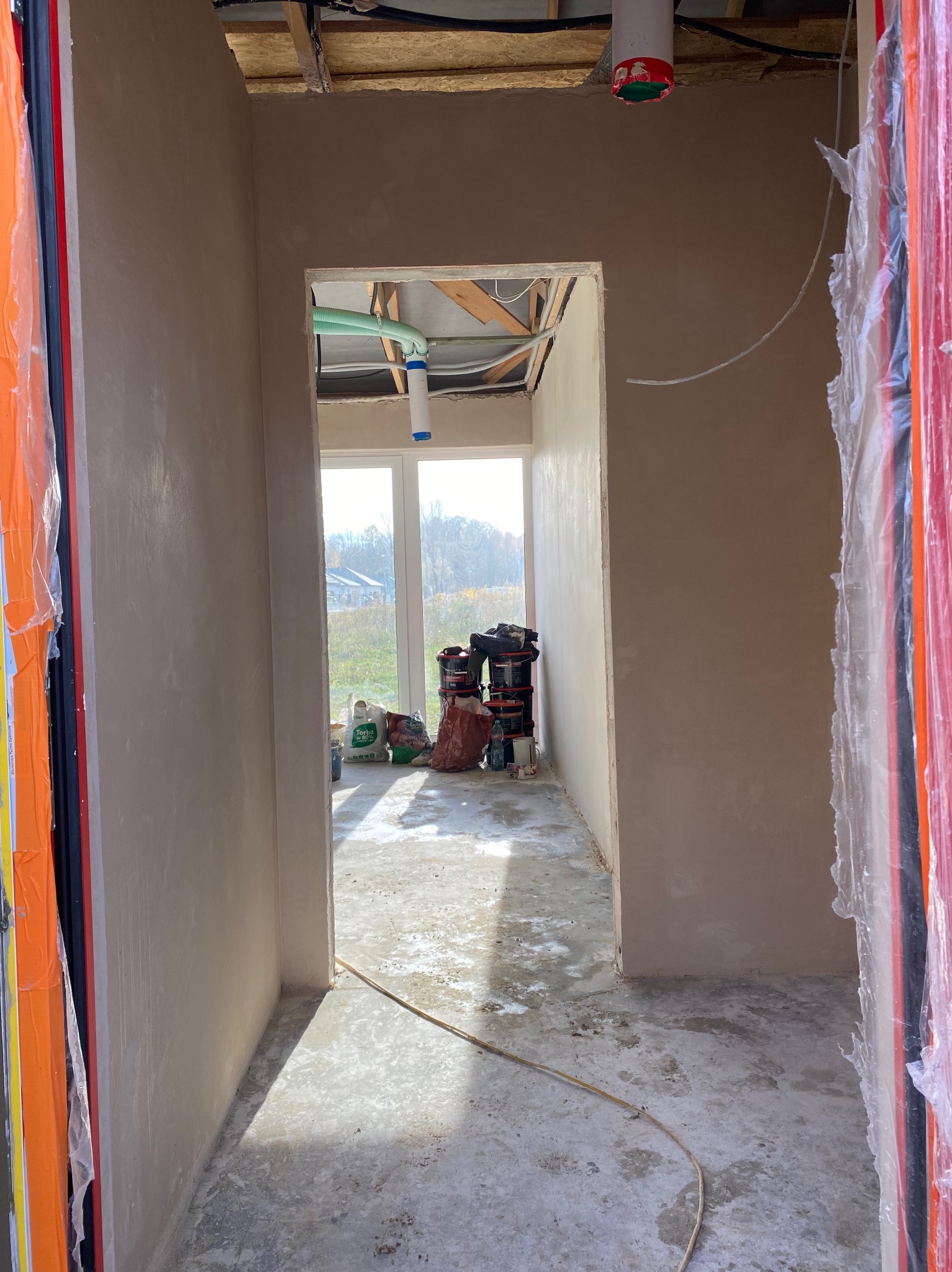 Wnętrze w trakcie prac wykończeniowych: ściany pokryte świeżym tynkiem, widoczne worki z materiałami budowlanymi i instalacje pod sufitem. Surowe, jasne pomieszczenie.