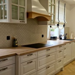 Adastol - Producent mebli na wymiar - Jasna kuchnia w stylu klasycznym: białe fronty szafek z frezowaniami i ozdobnymi uchwytami, drewniany blat, mozaika na ścianie i okno z roletami rzymskimi.