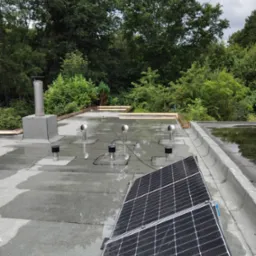 Płaski dach pokryty papą z elementami wentylacji, kominem i częściowo zamontowanymi panelami solarnymi. Widoczne kałuże po deszczu i otoczenie drzew.